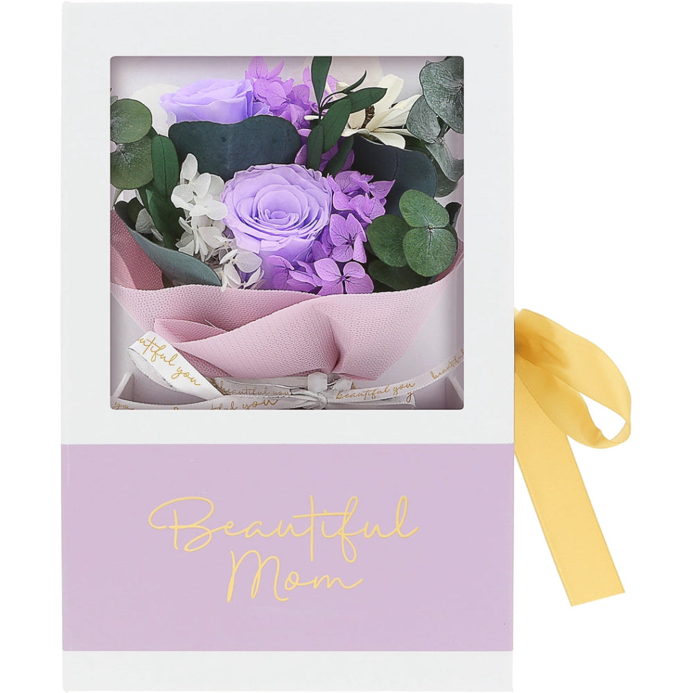 A floral gift box for Mom featuring a bouquet of purple and white flowers with green eucalyptus leaves inside a clear window, wrapped in pink paper with a white ribbon. The box has a lavender lower section with gold script reading 'Beautiful Mom' and a yellow ribbon tied to the side.