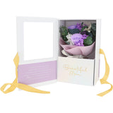 A white floral gift box with a clear window on the left panel and a purple flower arrangement inside on the right. The box is decorated with a lavender message card, gold 'Beautiful Mom' script, and yellow ribbon ties, shown on a white background.